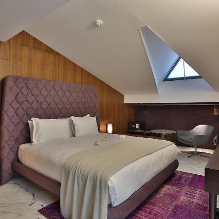 Dosso Dossi Hotels & Spa Downtown Istanbul Ngoại thất bức ảnh The photo shows a stylish hotel room with a modern design. It features a large bed with a tufted purple headboard, complemented by white bedding and a light gray throw. The walls are made of wooden panels, adding warmth to the space. To the side, the