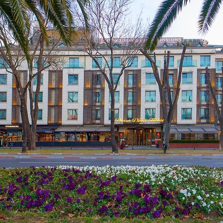 Dosso Dossi Hotels & Spa Downtown Istanbul Ngoại thất bức ảnh The photo shows a building with a modern design, featuring a facade with large windows and a mix of materials. In the foreground, there are colorful flower beds with purple and white flowers. The scene is framed by tall trees, and the overall setting