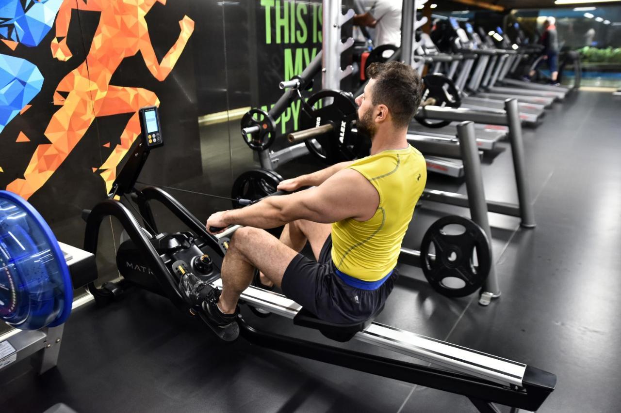 Dosso Dossi Hotels & Spa Downtown Istanbul Ngoại thất bức ảnh The photo shows a man exercising on a rowing machine in a gym. He is wearing a yellow tank top and black shorts. The background features an exercise-themed mural with colorful graphics of a person in motion. Various gym equipment, including treadmill