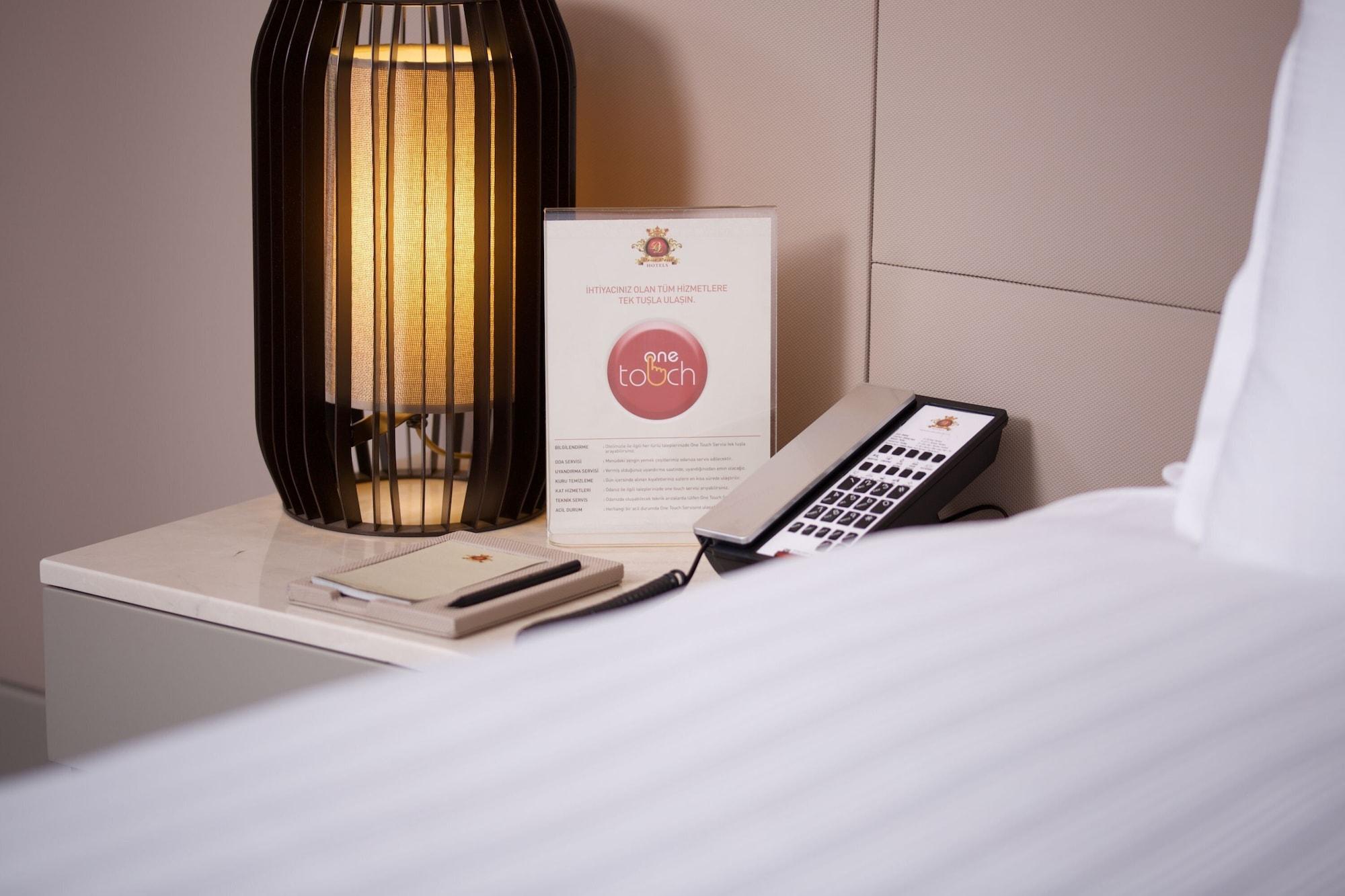 Dosso Dossi Hotels & Spa Downtown Istanbul Ngoại thất bức ảnh The photo shows a bedside setup in a room. There is a stylish lamp with a warm light on a table, alongside a menu or information card that features a logo or brand name. A remote control is also placed on the table, suggesting it might be used for va