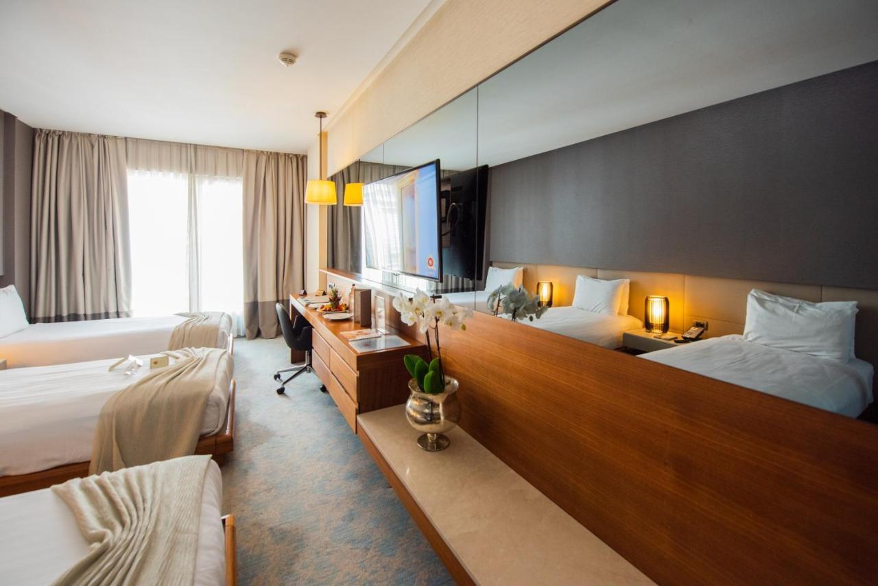 Dosso Dossi Hotels & Spa Downtown Istanbul Ngoại thất bức ảnh The photo shows a modern hotel room featuring two beds with light-colored bedding. There is a large mirror above a wooden dresser, which holds some decorative items, including a small plant. A TV is mounted on the wall, and there is a workspace with 