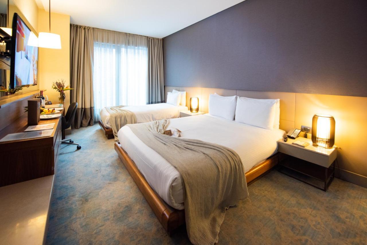 Dosso Dossi Hotels & Spa Downtown Istanbul Ngoại thất bức ảnh The photo shows a modern hotel room with two beds. One bed is covered with a light-colored blanket, and there are white pillows on both beds. The walls are painted in a soft, neutral tone, while one wall is a darker shade. There are large windows wit