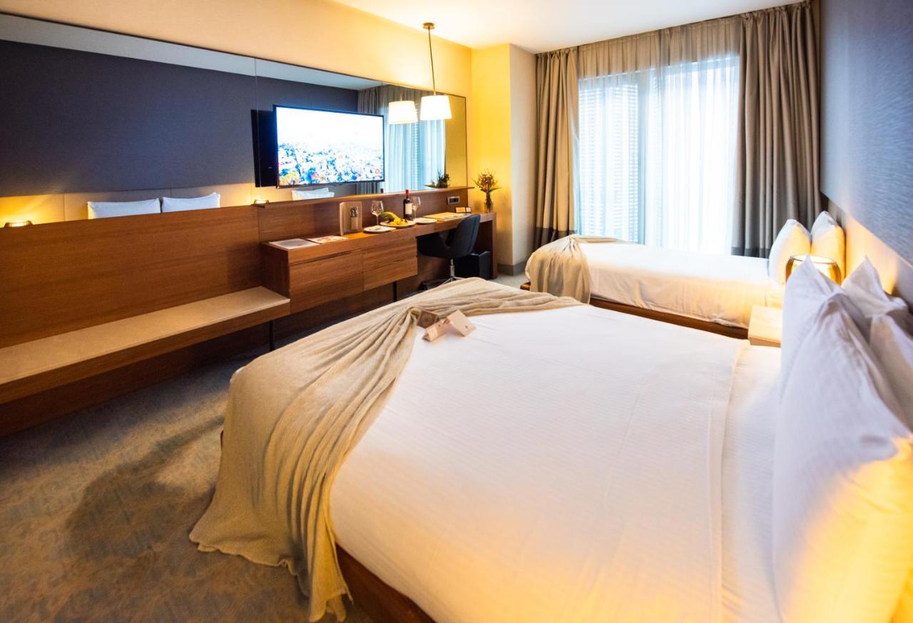 Dosso Dossi Hotels & Spa Downtown Istanbul Ngoại thất bức ảnh The photo shows a modern hotel room featuring two beds with white linens and a decorative throw blanket. There's a wooden desk and a chair alongside a large flat-screen TV mounted on the wall. The room has large windows with curtains, allowing natura