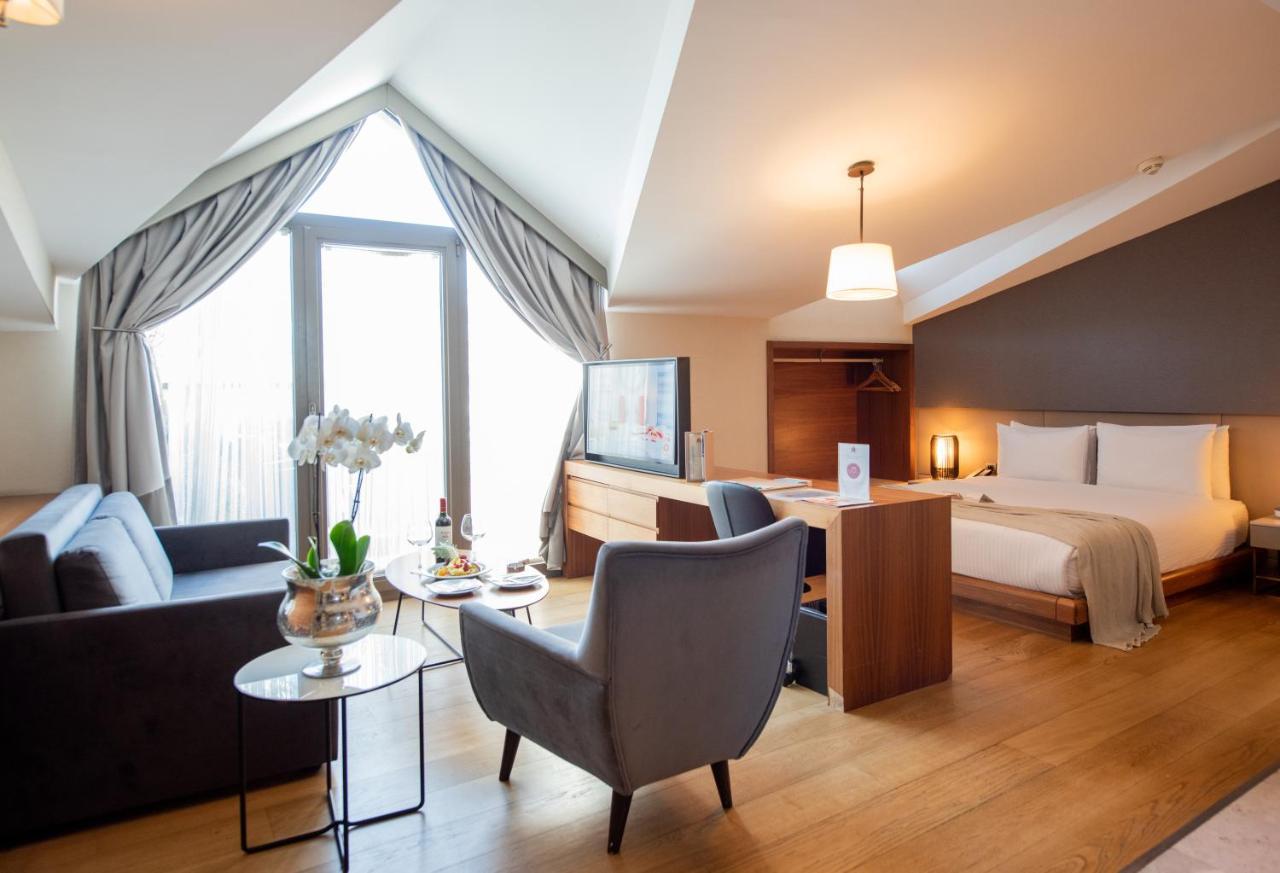 Dosso Dossi Hotels & Spa Downtown Istanbul Ngoại thất bức ảnh The photo shows a stylish hotel room with a modern design. It features a comfortable bed in one area, complemented by a cozy seating area with a sofa and a chair. There is a small round table with a vase of flowers and some snacks. A flat-screen TV i