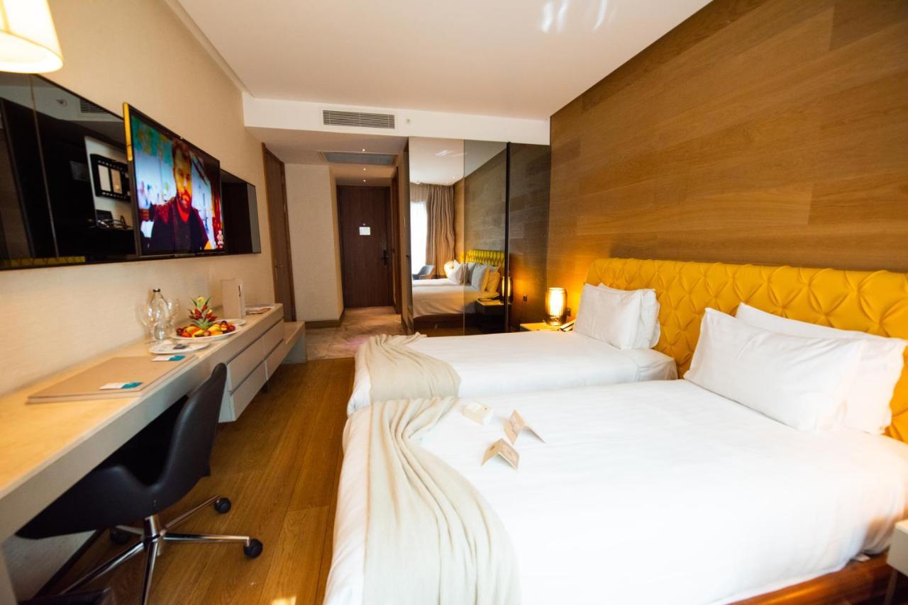 Dosso Dossi Hotels & Spa Downtown Istanbul Ngoại thất bức ảnh The photo shows a modern hotel room featuring two single beds with white linens. There is a stylish yellow accent wall behind the beds. A flat-screen television is mounted on the wall opposite the beds, and a sleek desk and chair are positioned next 