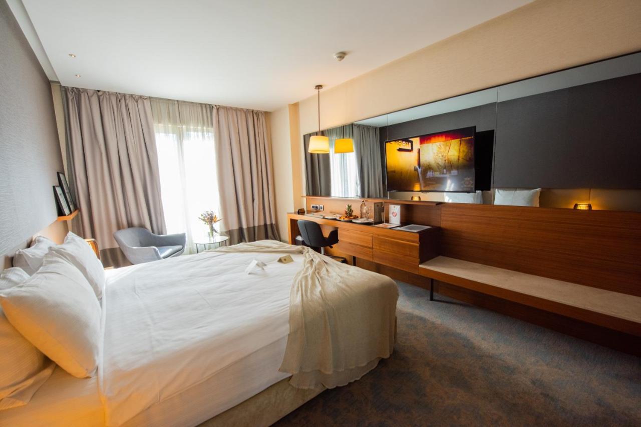 Dosso Dossi Hotels & Spa Downtown Istanbul Ngoại thất bức ảnh The photo shows a modern hotel room featuring a large bed with white linens, set against a soft-toned wall. There is a stylish gray armchair in one corner and a sleek wooden desk with a chair. The room is well-lit, with large windows covered by light