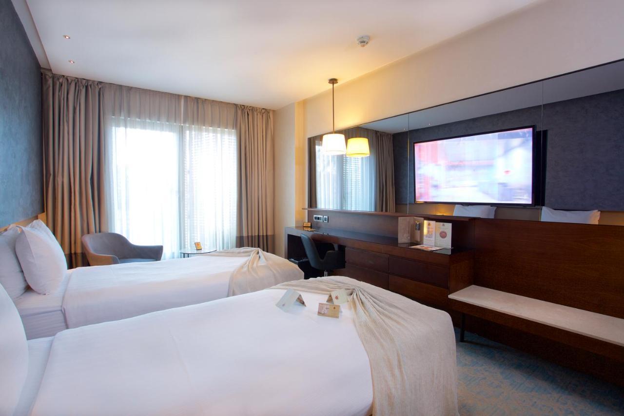 Dosso Dossi Hotels & Spa Downtown Istanbul Ngoại thất bức ảnh The photo shows a hotel room featuring two twin beds with white linens. There is a large window with curtains that allows natural light to enter the room. A wooden cabinet is present, along with a flat-screen television mounted on the wall. Additiona