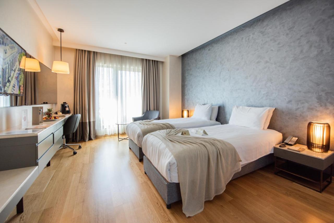Dosso Dossi Hotels & Spa Downtown Istanbul Ngoại thất bức ảnh The photo shows a modern hotel room featuring two single beds with white linens. The room has a muted color palette, including a gray wall and light wooden flooring. Large windows allow natural light to flood the space, and there are sheer curtains f