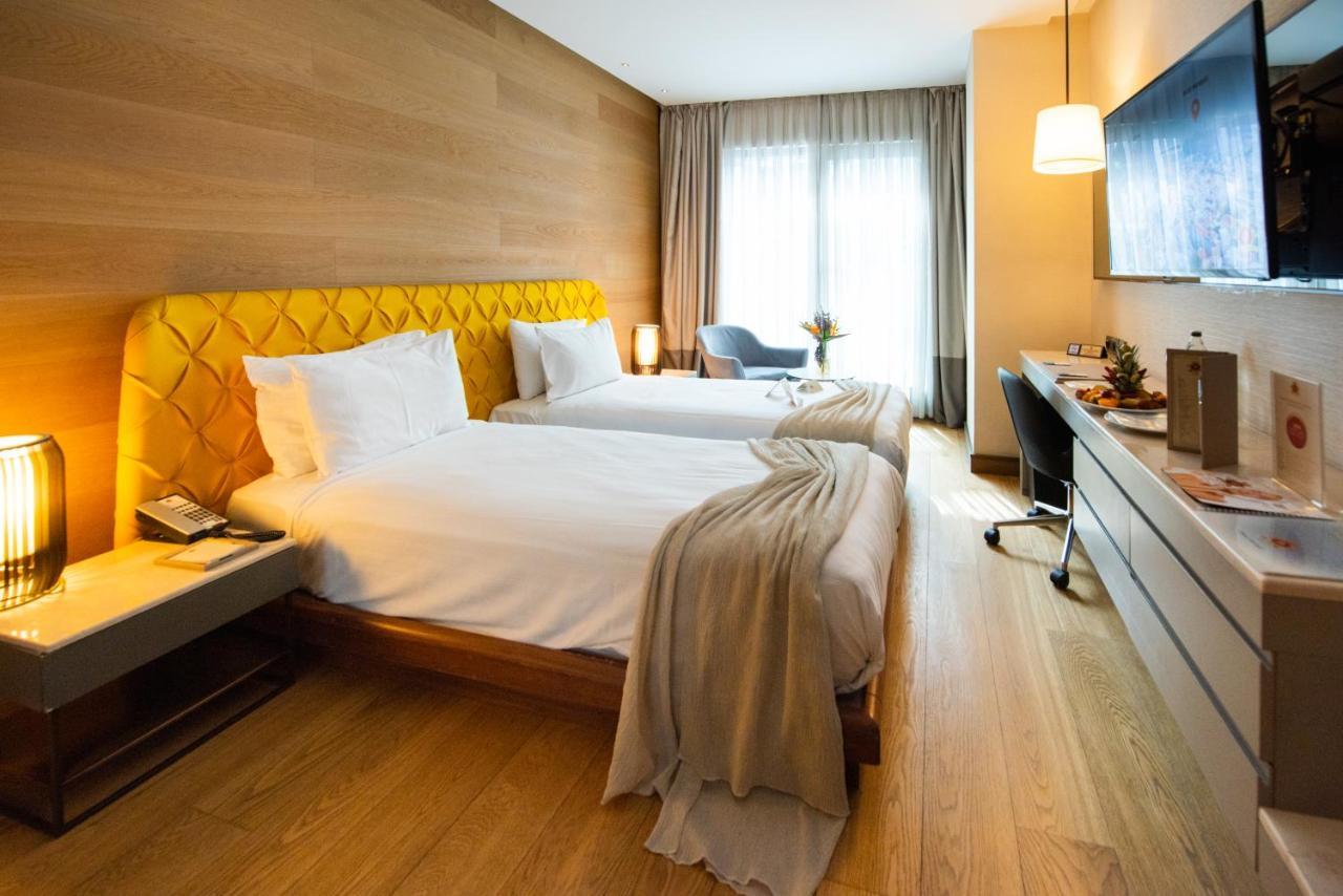 Dosso Dossi Hotels & Spa Downtown Istanbul Ngoại thất bức ảnh The photo shows a stylish hotel room featuring two beds with white linens and a unique yellow headboard. The room has wooden walls and a polished wooden floor. There are large windows with sheer curtains allowing natural light to flood in. A small se