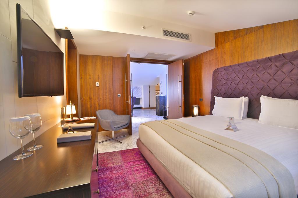 Dosso Dossi Hotels & Spa Downtown Istanbul Ngoại thất bức ảnh The photo depicts a modern hotel room. It features a large bed with white linens and a decorative headboard in a rich purple hue. The room has wooden paneling on the walls, adding warmth to the space. There's a flat-screen TV mounted on the wall oppo
