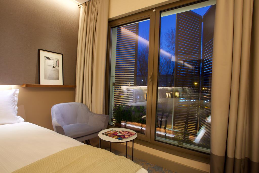Dosso Dossi Hotels & Spa Downtown Istanbul Ngoại thất bức ảnh The photo depicts a modern hotel room. On the left side, there is a neatly made bed with a light-colored blanket. Next to the bed, there's a small round table with some magazines on top. A cozy chair in a light shade is positioned nearby. The main fe